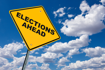 A tilted yellow road sign with black text reading "Elections Ahead" stands against a backdrop of a blue sky filled with white clouds. The sign not only suggests upcoming elections but also hints at opportunities for local employment.