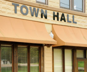 A building with a brick facade has a sign that reads "Town Hall" above a row of windows covered by orange awnings. The lower part of this local government building has several windows with horizontal blinds partially drawn.