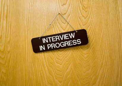 A small rectangular black sign with white text hangs on a wooden door, reading "INTERVIEW IN PROGRESS". The interview sign is suspended by a thin metal chain.