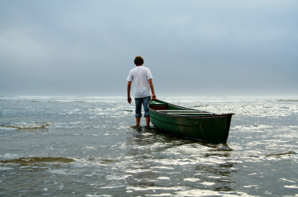 A person wearing a white shirt and rolled-up jeans stands in shallow water, holding onto a green canoe. The sky is overcast, and the sea extends to the horizon, blending with the cloudy sky. One wonders what adventures the future has in store for you?