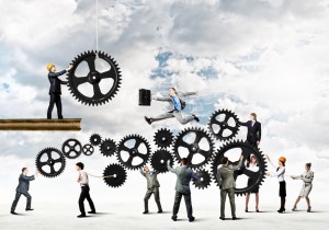 A group of people in business attire interact with large interlocking gears against a cloudy sky backdrop. One person in a hard hat operates a suspended gear, while another jumps over gears holding a briefcase. Others push or pull the gears, symbolizing the effort of retaining employment amidst challenges.