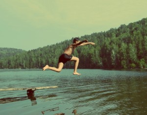 A person in swim trunks jumps off a wooden platform into a lake surrounded by lush, green trees on a clear day. The scene captures a mid-air moment of exhilaration, with the forested shoreline and calm waters in the background, reminding us of the importance of public safety while enjoying nature.