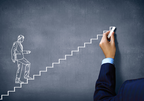 A hand in a suit is drawing white steps on a dark chalkboard. A sketch of a person in a business suit is shown stepping upward on the drawn stairs, symbolizing career progress and achievement.