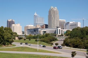 A cityscape of Raleigh, North Carolina on a clear day, featuring several tall buildings, including the distinctive PNC Plaza and Two Hannover Square. Cars are driving on a roadway in the foreground, surrounded by green trees and grassy areas.