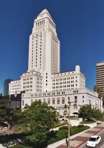 A tall, white, multi-story building with a prominent tower rises against a clear blue sky. The building, surrounded by greenery, features the American and Californian flags on one side, and a few vehicles are visible on the road in front.