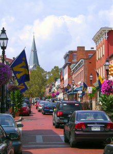 A lively street scene featuring cars parked along a brick-paved road lined with colorful buildings. Flags and hanging flower baskets adorn lamp posts. A church steeple rises in the background under a partly cloudy sky.