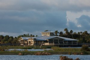 Rectangular building with large windows and a metal roof situated in a marshy area with water in the foreground. A boardwalk extends from the building. The sky is partially cloudy, and palm trees and other vegetation surround the structure.