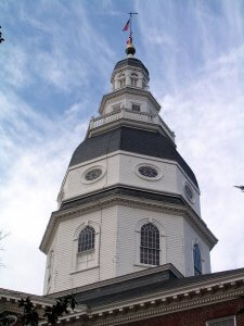 A historic white wooden dome with architectural details and multiple tiers stands tall against a cloudy sky. An American flag is flying from a pole at the dome's peak. The structure is part of a larger red brick building visible at the bottom of the image.