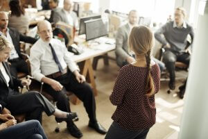 A group of people in business attire are seated around an office, engaged in discussion. A woman with a braided ponytail stands facing them, addressing concerns about gender bias, while open laptops and paperwork are visible on the desks. Natural light filters through the windows, illuminating the room.