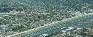 An aerial view of an airport runway adjacent to a densely populated neighborhood. Several small aircraft are parked on the tarmac. Residential homes and green spaces extend beyond the airport, with streets visibly dividing the community.