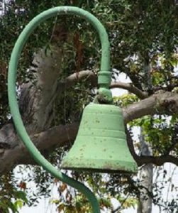 A green metal bell hangs from a curved pole attached to a tree branch with lush green leaves in the background. The bell and pole show signs of wear and rust.