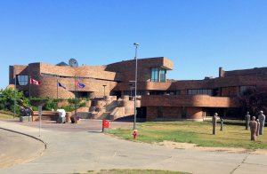 A modern brick building with multiple levels and large windows, featuring a central staircase and entrance. It has flagpoles with various flags, several sculptures of humanoid figures, a green lawn, and a clear blue sky in the background.