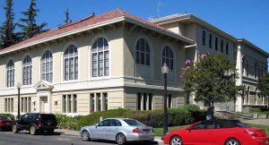 A large, beige neoclassical-style building with arched windows and a red-tiled roof is shown on a sunny day. Several cars are parked along the street in front. A lamp post and trees are visible nearby. The sky is clear and blue.