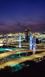 An illuminated urban landscape at night, featuring a network of intersecting highways. Tall, colorfully lit pillars line the roads, and a brightly lit city with various buildings is visible in the background under a clear sky.