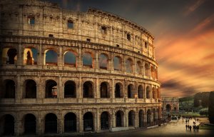 The Colosseum in Rome at sunset, with its ancient stone arches illuminated by the glow of the setting sun. A group of tourists, guided by a leadership tour, is seen near the base, while the sky is painted in hues of orange, pink, and purple.