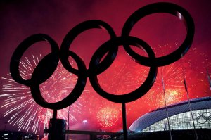 Giant Olympic rings are silhouetted against a vivid red sky illuminated by a colorful fireworks display over Los Angeles. The bright bursts of fireworks light up the scene, with a modern stadium building visible in the background.