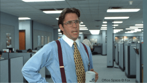 A man wearing glasses and a blue dress shirt with burgundy suspenders stands in an office holding a white mug. With a serious expression, he surveys the large room filled with cubicles, embodying the essence of bosses everywhere. Text in the bottom right corner reads "Office Space © Fox.