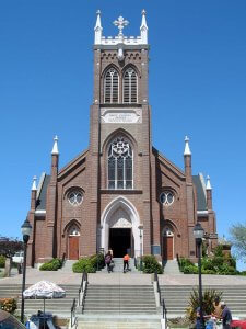 A tall brick church with a large central bell tower stands under a clear blue sky. The church features pointed arch windows and a detailed entrance. Two people are ascending the stairs to the entrance. The foreground has street lamps, a few plants, and visitors.