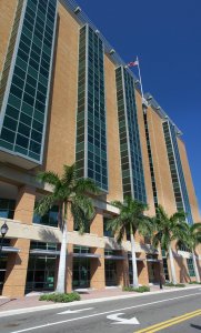 A tall modern building with large glass windows and a brick facade stands under a clear blue sky. Rows of tall palm trees line the street in front of the building. An American flag is visible atop the building.