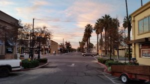 A quiet urban street at dawn with palm trees lining the sidewalk. Buildings and shops flank the street, including a beige building on the right with a "RENT" sign. A white truck and a brown trailer are parked at the foreground. The sky is painted with pink and blue hues.