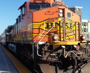 A bright orange BNSF locomotive is stationed on a track next to a platform. The front of the engine is prominently displayed, showing its distinctive design and colors. A passenger train is seen in the background on the adjoining track. The scene is set in a daylight.