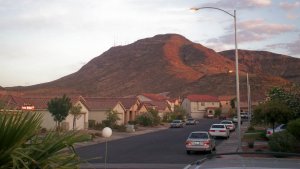 A suburban neighborhood at dusk with parked cars and houses with tiled roofs. A large hill or small mountain, partially lit by the setting sun, rises in the background. Several streetlights line the street, and a palm tree is visible in the foreground.