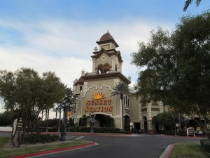 A wide-angle view of the Sunset Station hotel and casino. The building has a clock tower with a peaked roof and ornate design. Tall palm trees line the entrance and the logo with a sunburst design is prominently displayed above the main entrance. The sky is partly cloudy.