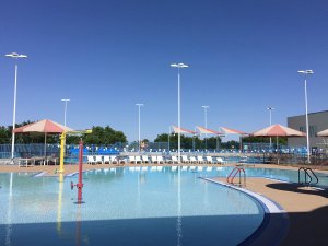 A large outdoor swimming pool with crystal-clear water under a clear blue sky. The pool area features several shaded sitting areas with lounge chairs, bright red and yellow water play structures, and tall light posts surrounding the pool.