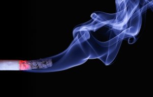 A lit cigarette with a burning tip is shown against a black background. Gray smoke rises and swirls gracefully from the burning end, creating intricate patterns in the air. The image captures the dynamic and ephemeral nature of the smoke, serving as a stark reminder of its impact on wellbeing.