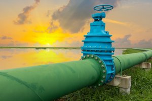 A large green industrial pipeline, part of the intricate water systems, with a blue valve runs through a grassy area and stretches towards the horizon, where the sun is setting over a body of water. The sky is filled with dramatic clouds, glowing in hues of orange and yellow.