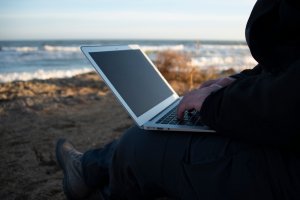 Person sitting on the ground by the ocean, working remotely on a laptop. The beach is visible in the background with waves gently rolling in. The person is dressed in dark clothing, and the scene suggests a peaceful, outdoor workspace.