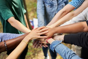 A group of diverse individuals extending their arms toward the center, stacking their hands on top of each other in a display of unity and humanitarian teamwork. The background shows a blurred outdoor setting.