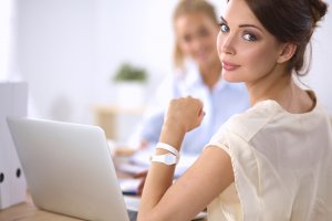 A woman with brown hair is sitting at a desk, looking back over her shoulder. She is wearing a beige blouse and a white wristwatch. In front of her is an open laptop, hinting at her busy career. Another person is sitting in the background, slightly out of focus.