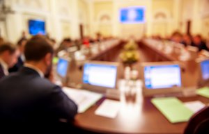 A blurred image showing people seated at a large, long table in a formal meeting room. Computer screens and documents are visible on the table, with a central display screen in the background. The setting suggests a government conference, meeting, or official discussion.