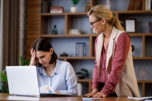 A young woman sitting at a desk with a laptop holds her head in frustration while looking at papers. An older woman stands next to her, leaning over with a concerned expression. The background features various workplace decorations on a wooden bookshelf.