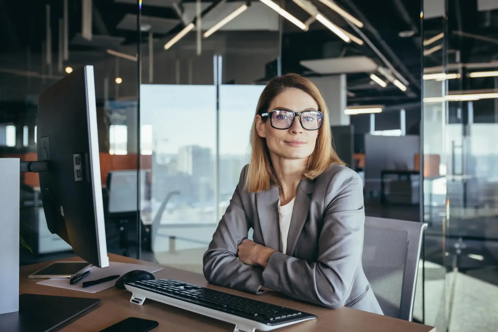 A woman with shoulder-length hair and glasses sits confidently at a desk in a modern office. She is wearing a gray blazer and looking directly at the camera. A computer monitor, keyboard, and office furnishings are visible around her.