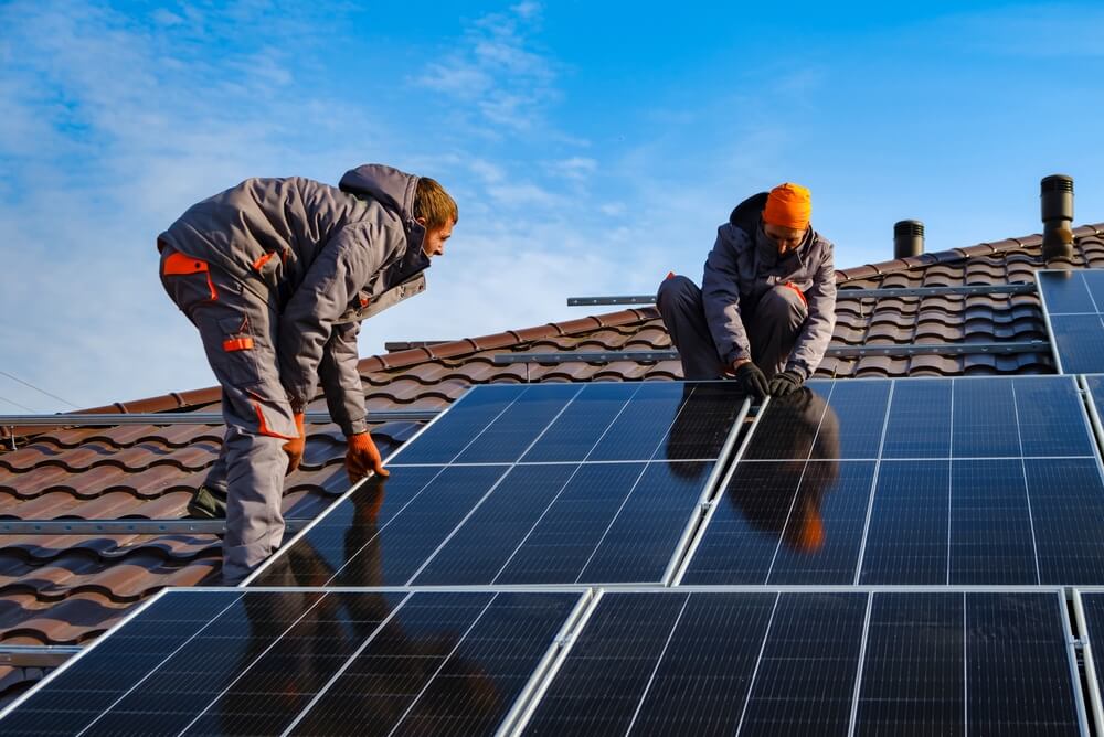 Two workers in gray and orange uniforms, part of a government initiative, are installing solar panels on a tiled rooftop under a clear blue sky. They are carefully positioning the panels, with one standing and the other kneeling, ensuring proper alignment and attachment.