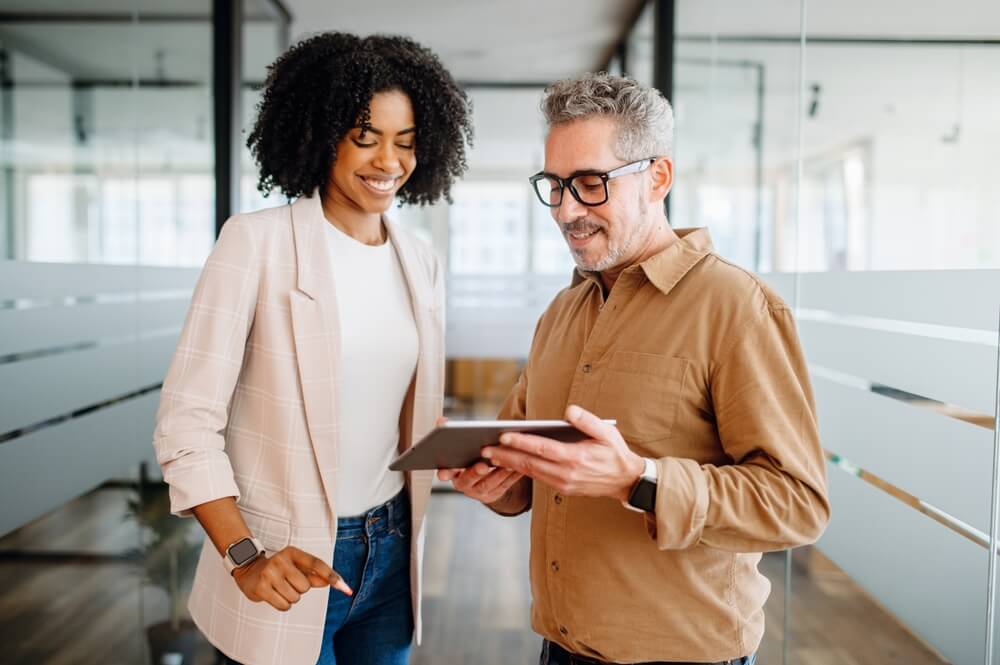 Two colleagues stand in a modern office with glass walls, engaged in a work-related discussion. One, a woman with curly hair, smiles while the other, a man with glasses and a beard, holds a tablet. Both appear to be analyzing or sharing information on the device.