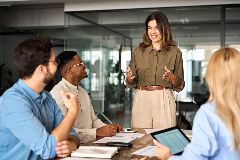 A woman exudes leadership as she stands and smiles while speaking to three colleagues seated around a table in a modern office. One person is taking notes in a notebook, another is using a laptop, and another has a tablet. They appear engaged in a collaborative discussion.