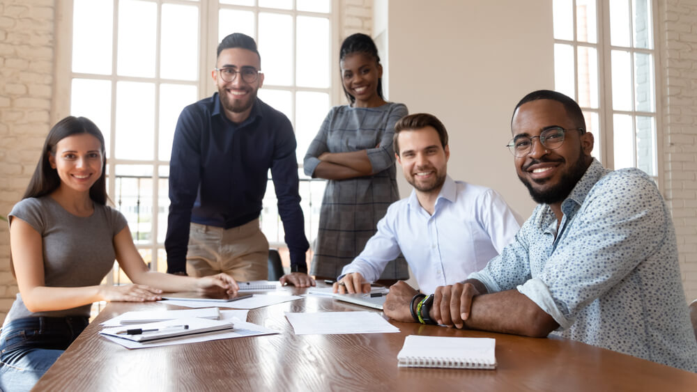 A group of five diverse professionals, three men and two women, are smiling and posing for a photo around a table with paperwork and pens. They appear to be in a well-lit government office space with large windows in the background.