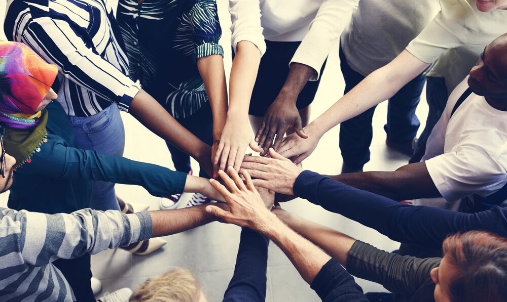 A diverse group of people from the public sector stands in a circle, extending their hands into the center to form a collective pile of hands, symbolizing unity and teamwork. The individuals wear various colorful clothing, and the background is blurred.