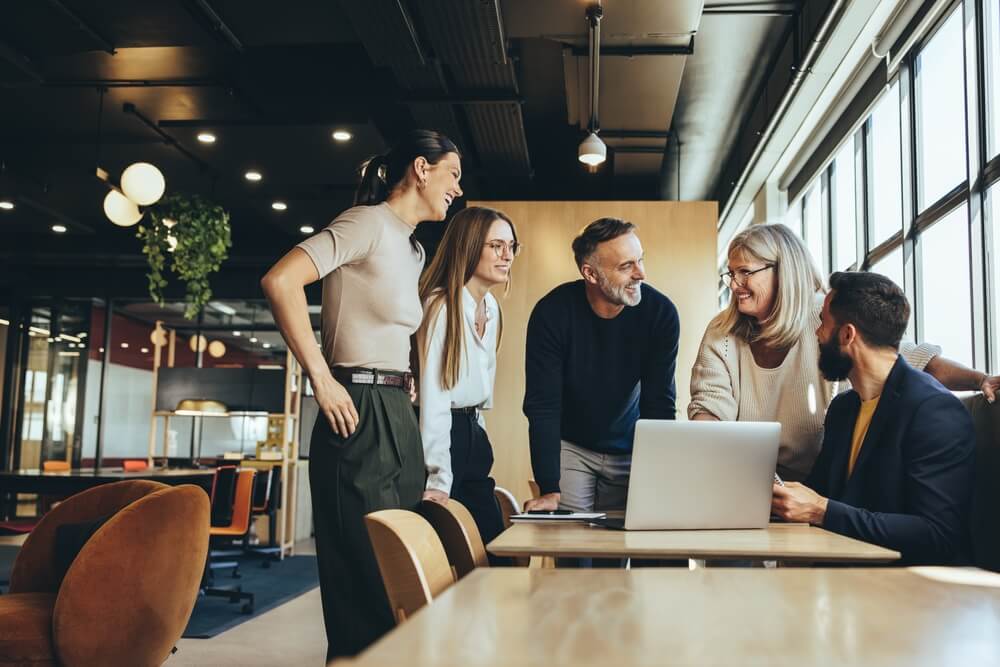 A group of five colleagues stands around a laptop in a modern office with large windows and warm lighting. They appear to be engaged in positive and animated work-related discussions. The office features contemporary furniture and potted plants, creating a welcoming atmosphere.