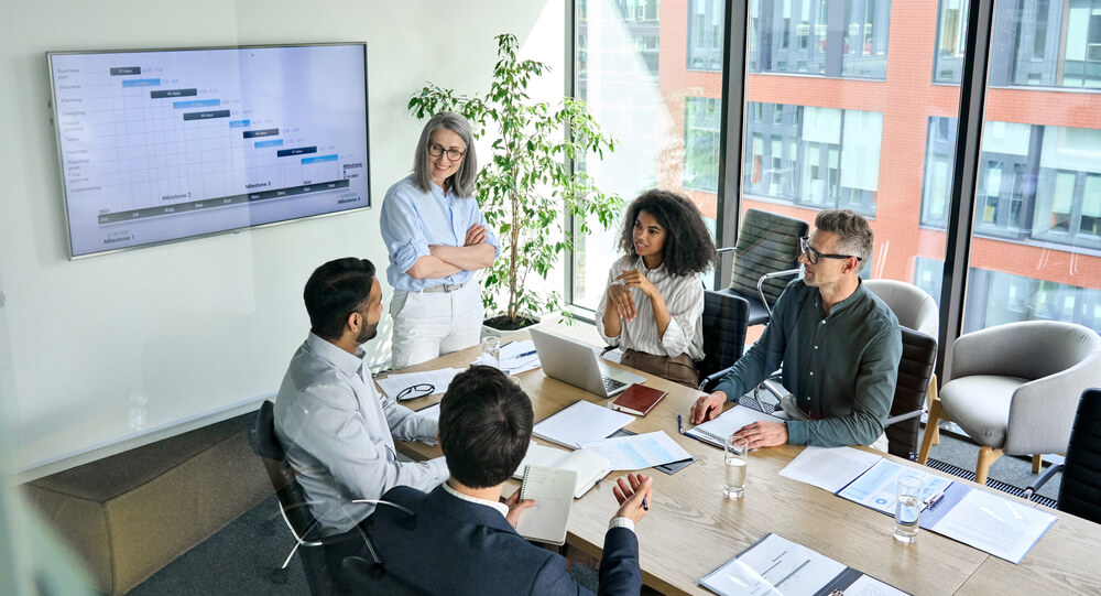 A group of five people are gathered around a conference table in a bright office with large windows. One person, demonstrating leadership, is standing and presenting a Gantt chart displayed on a wall screen. The seated individuals are attentively listening and taking notes.