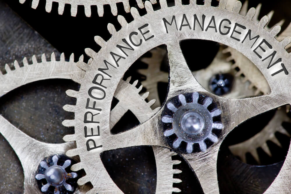 Close-up of several interlocking metal gears, with one large gear prominently displaying the words "PERFORMANCE MANAGEMENT" engraved on it. The intricate machinery symbolizes interconnected processes in public sector performance management systems.