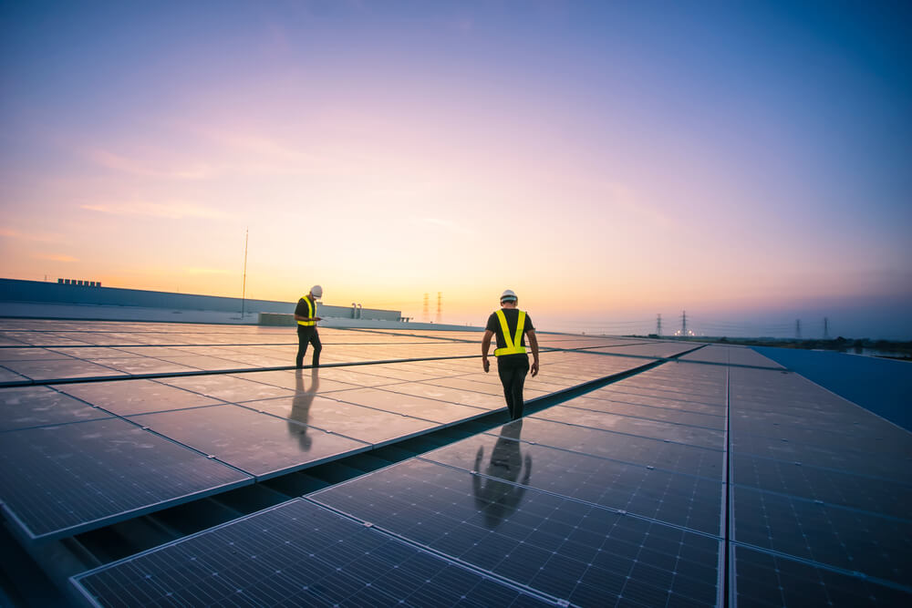 Two workers in safety vests and hard hats inspect a large array of solar panels on a rooftop at sunset, reflecting the setting sun's light. The gradient sky adds to the scene as electrical towers loom in the distance. This government-backed project exemplifies commitment to sustainable energy.