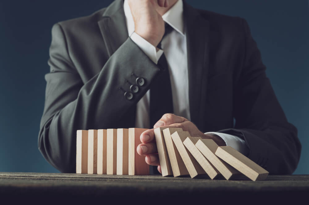 A person in a suit is seen from the chest down, with one hand preventing a row of wooden dominoes from toppling over. The scene suggests the act of preventing a potential chain reaction, much like managing issues within a workforce to maintain stability.