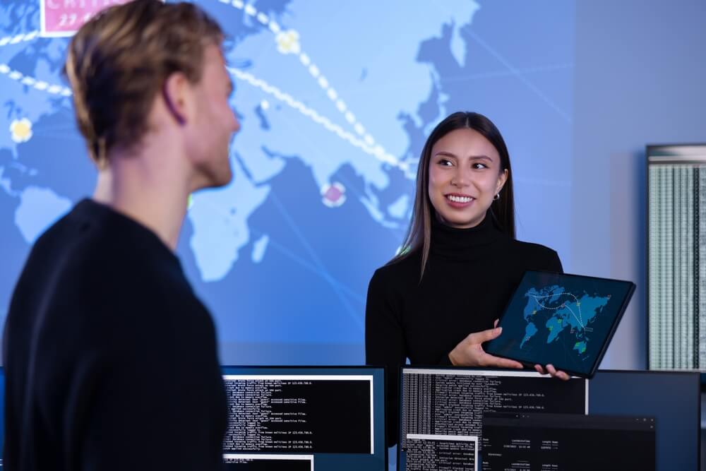 Two people in a tech environment. A woman holding a tablet displays a map with data points, and a man stands nearby looking at her and smiling. Multiple monitors with code and data are in the foreground, reflecting the shift towards remote working, and a larger world map is projected on the wall behind them.