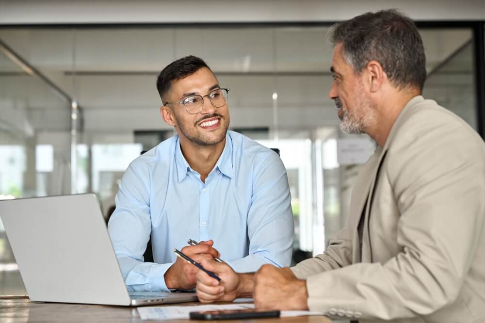 Two men sit at a desk in a modern office, engaged in conversation. The man on the left, wearing glasses and a light blue shirt, smiles while holding a pen, offering feedback. The man on the right, dressed in a beige blazer, listens attentively. A laptop is open on the desk.