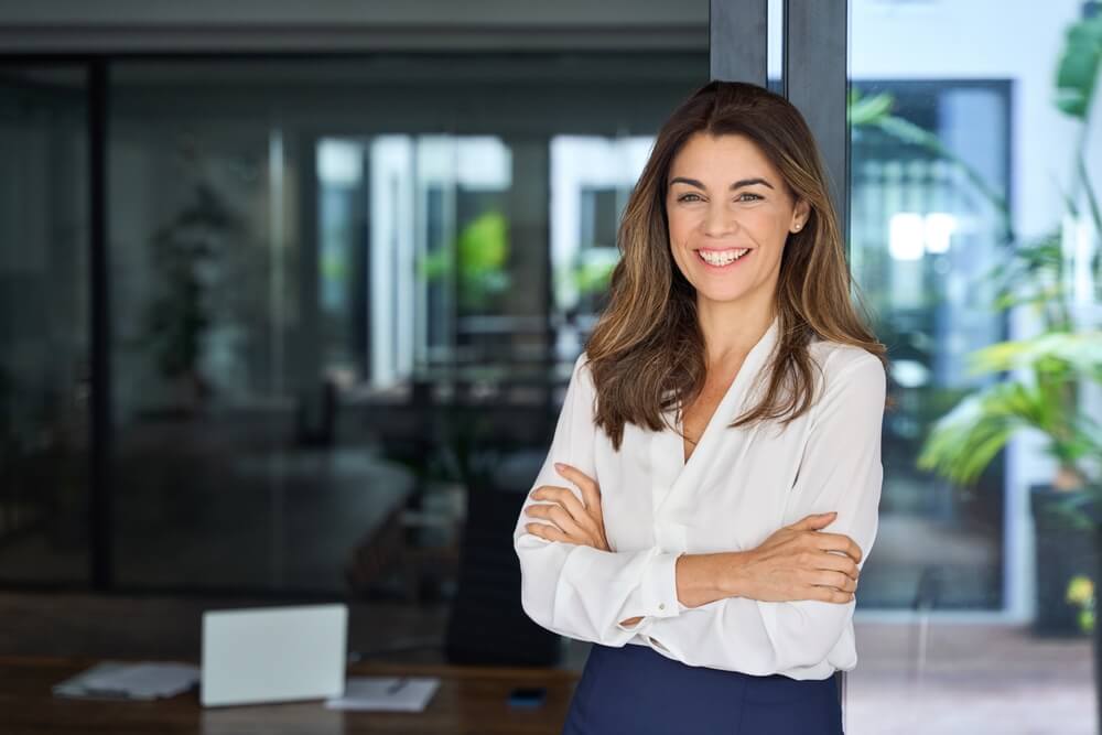 A confident woman with long brown hair and a white blouse stands with her arms crossed, smiling at the camera. Her intelligence shines through as she poses in a modern office setting with glass walls, a desk, and green plants in the background.