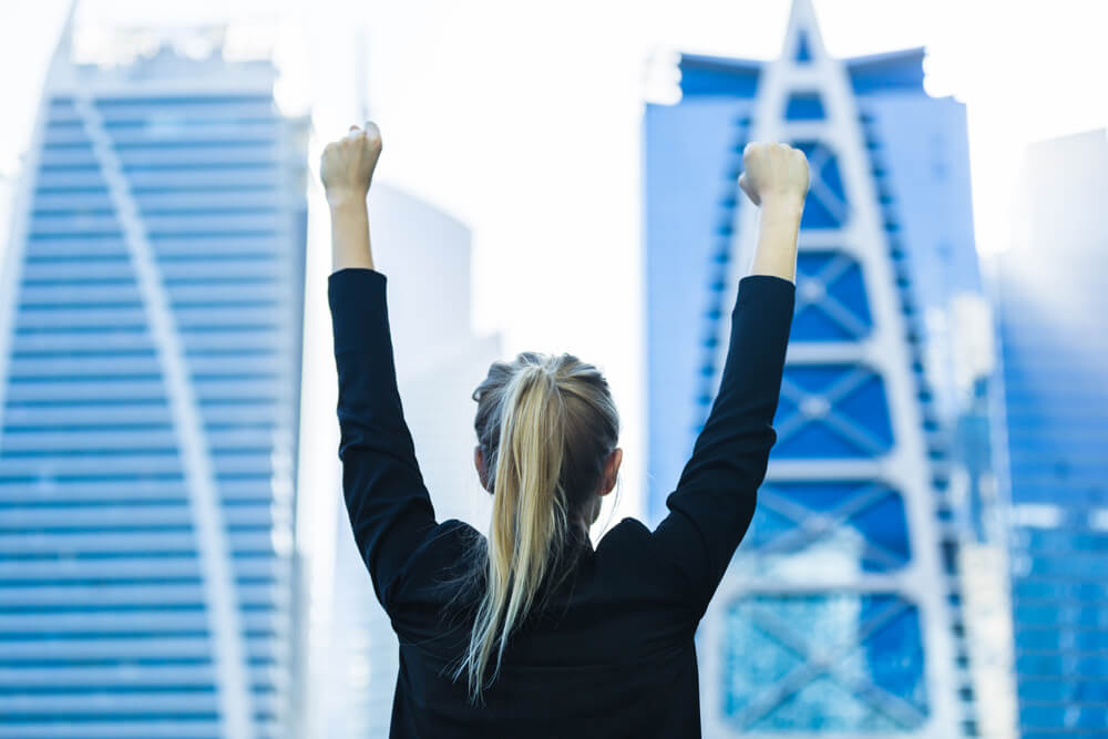 A person with long blonde hair, wearing a black jacket, stands with arms raised in a victory pose against a backdrop of modern skyscrapers in a cityscape. As they bask in the clear sky and the blue and white designs of the buildings, their mind is already journaling this triumphant moment.